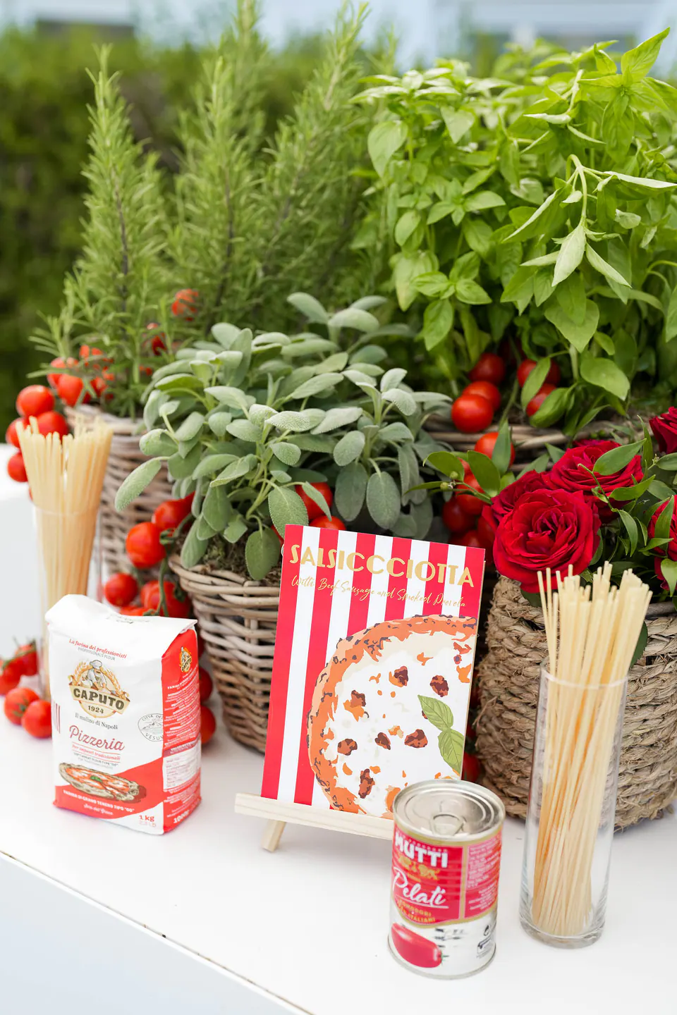 Table de la pizza party avec des paniers de basilic, sauge et romarin, des tomates cerises, des spaghettis et une boîte de tomates.