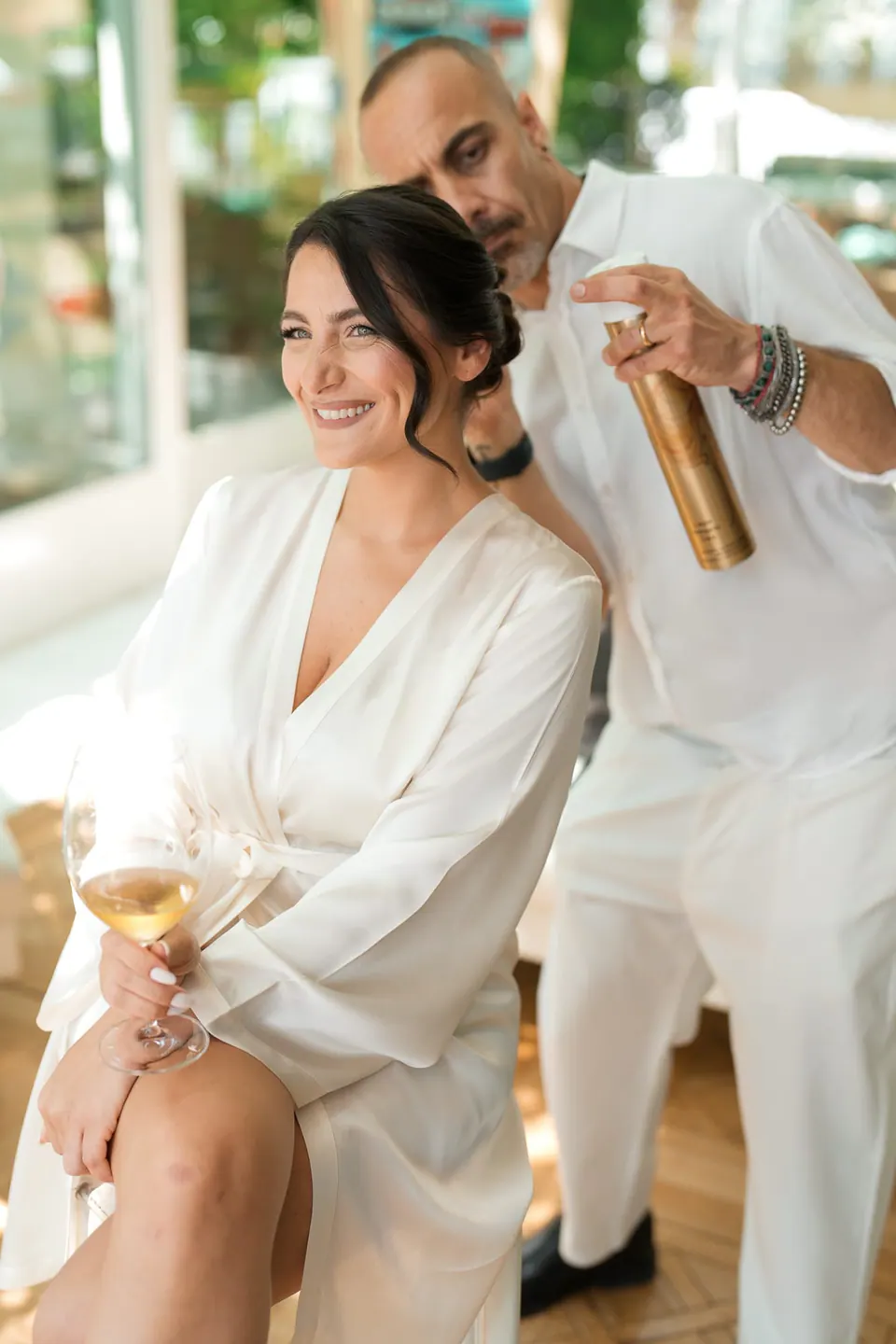 Die Friseurin sprüht Haarspray, die Braut hält ein Glas Wein.