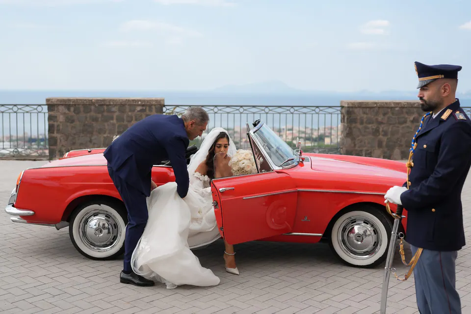Groom helping the bride out of the red convertible, a police officer in uniform standing by.