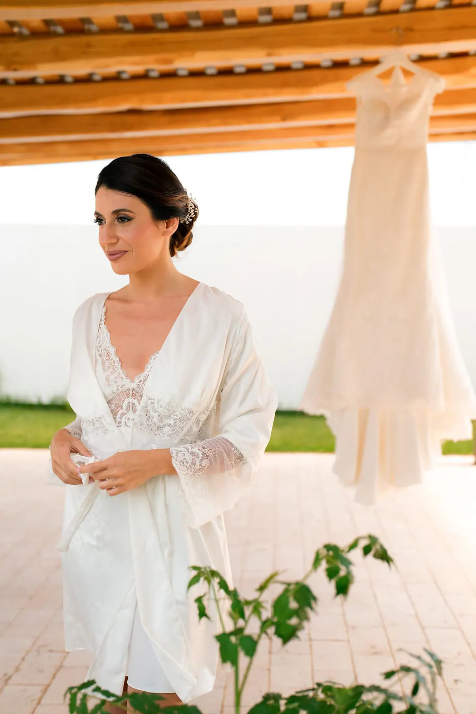 Bride in her robe under the pergola, her dress hanging behind her