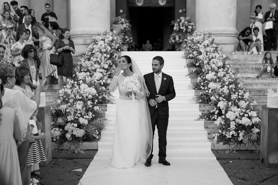 Black and white photo of the couple at the end of a white aisle lined with flowers among the columns.