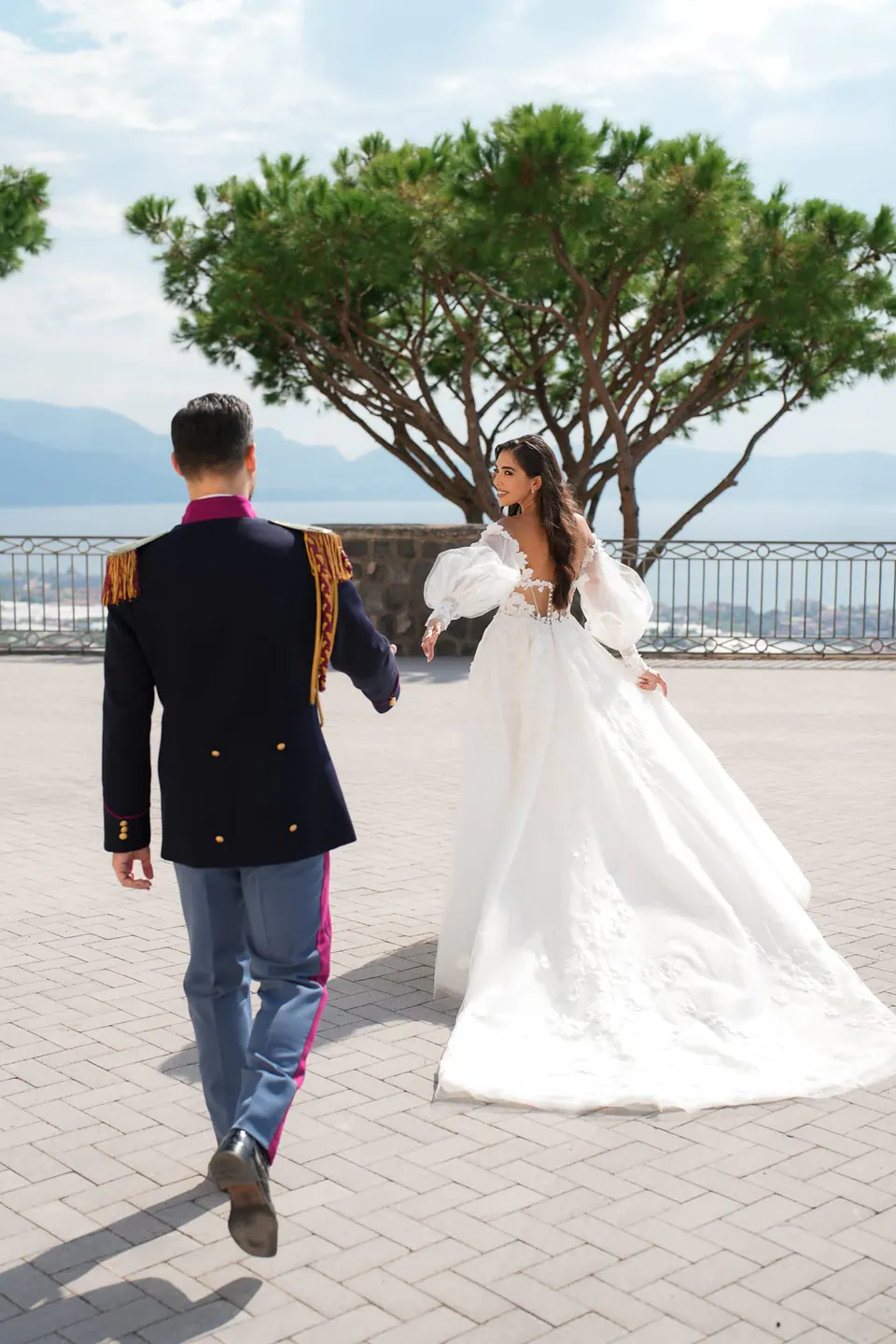 Groom in dress uniform walking toward the bride across the terrace beneath the umbrella pine.