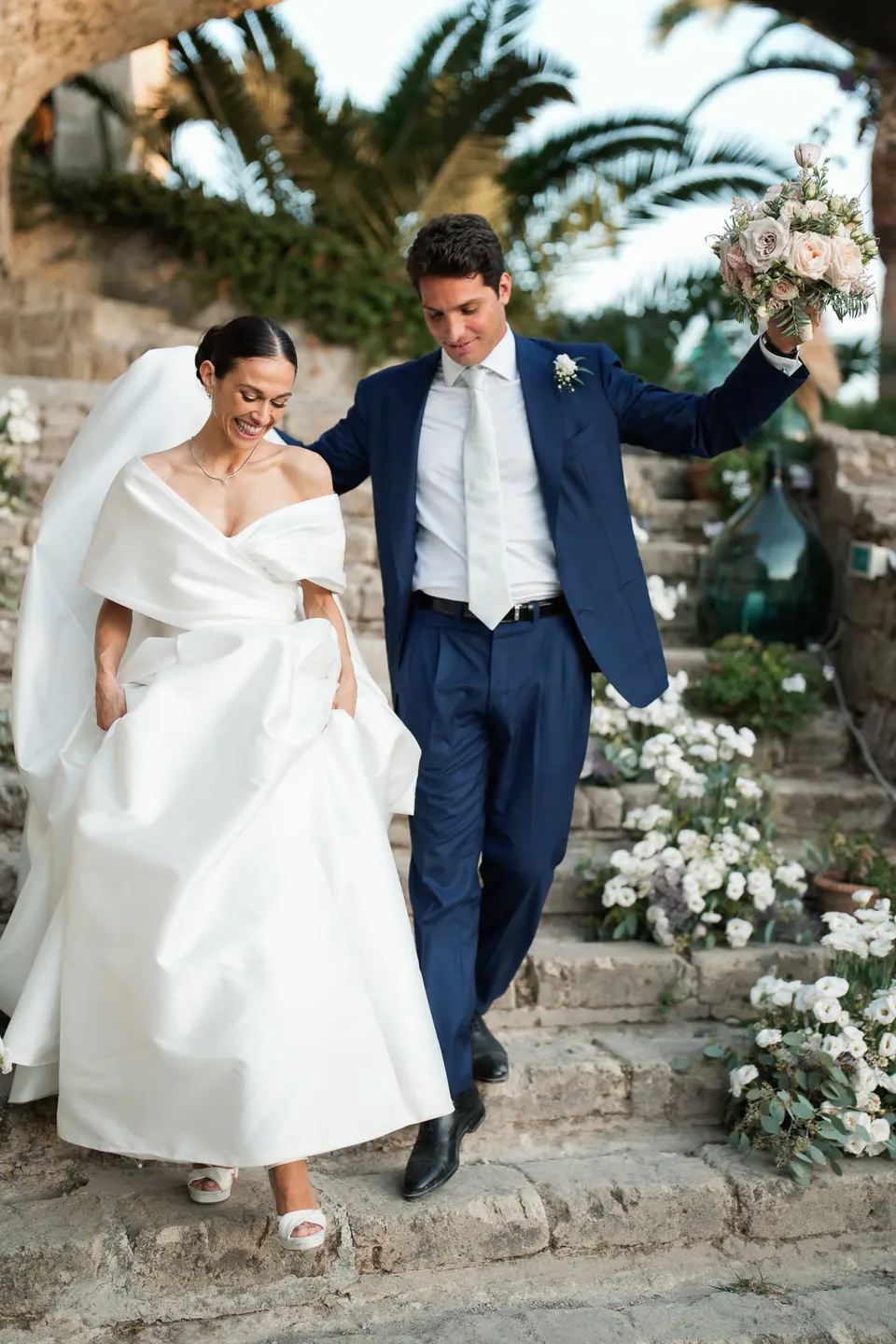 Sposi che scendono gradini del giardino ornati di fiori bianchi, lo sposo alza il braccio.