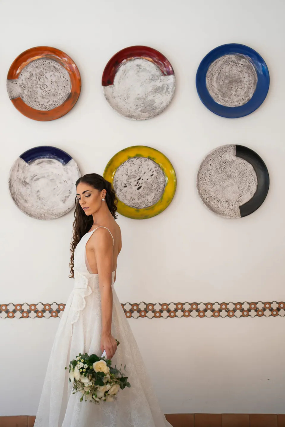 Bride with her bouquet beneath colourful ceramic plates on a white wall.