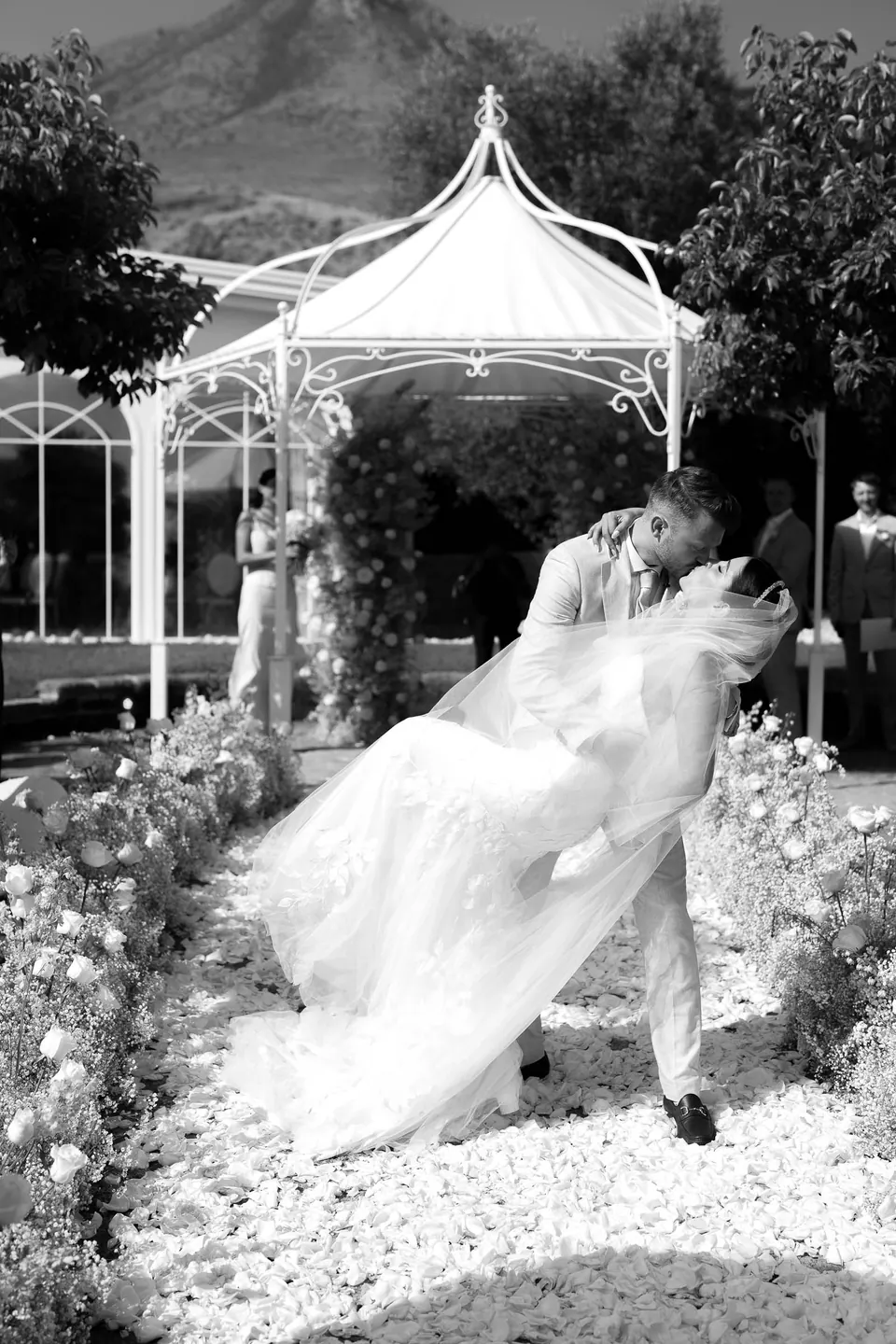 Le marié renverse la mariée pour un baiser devant le kiosque blanc du jardin, en noir et blanc.