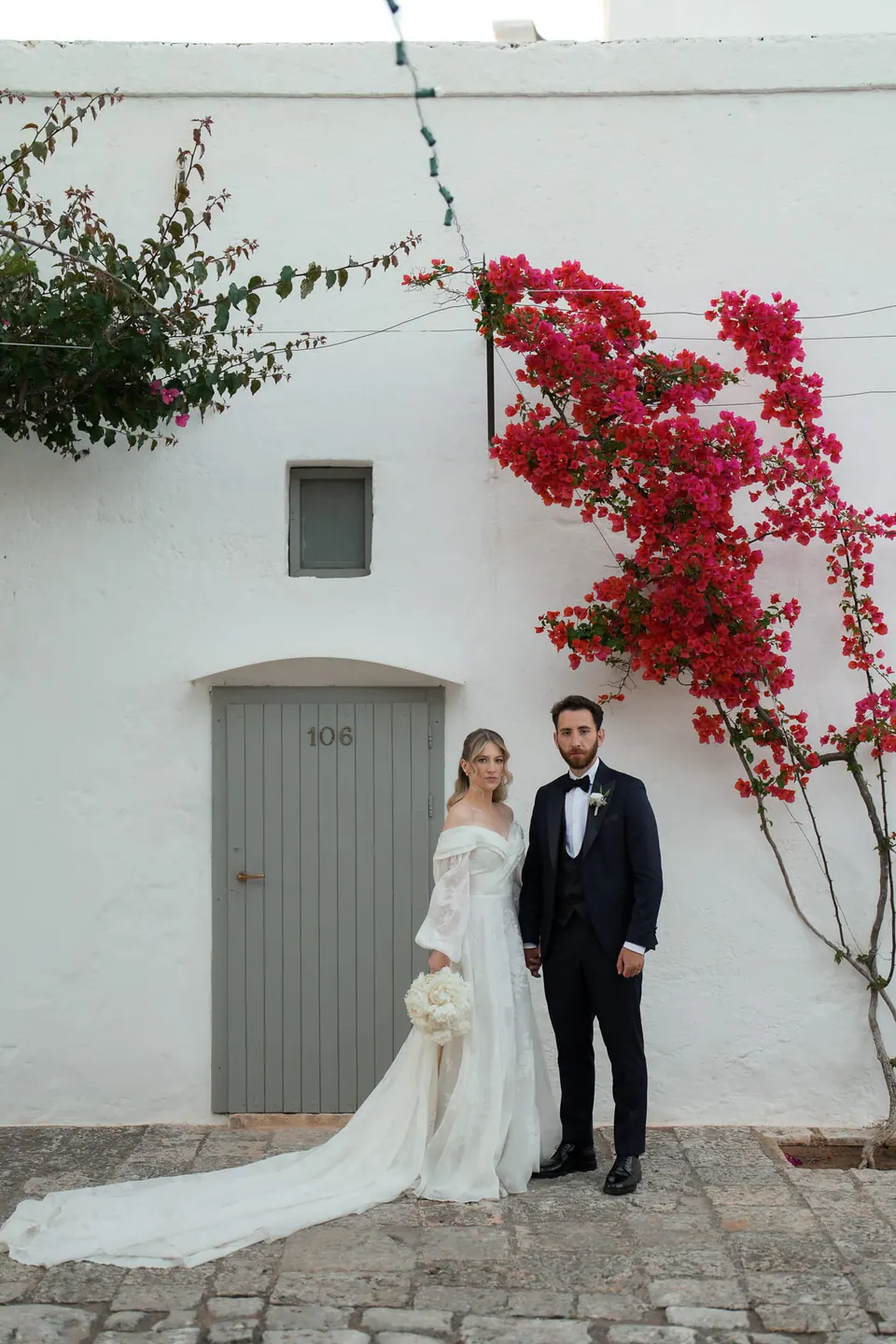 Los novios junto a una pared blanca bajo buganvillas rojas.