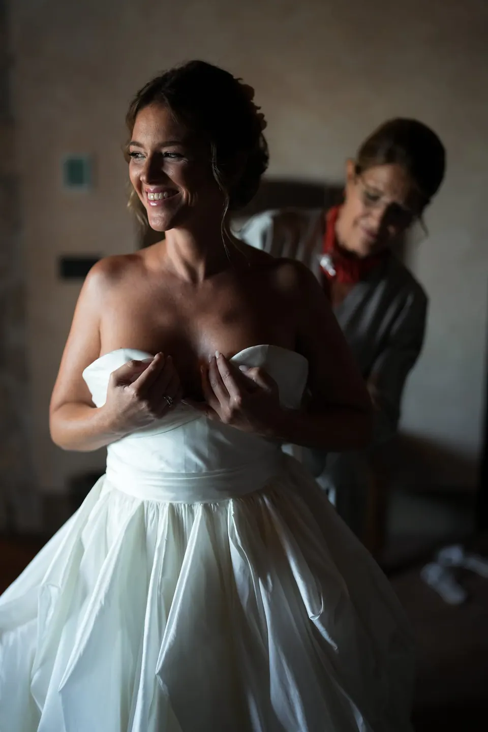 Sposa che ride mentre le allacciano l'abito, luce calda della finestra.