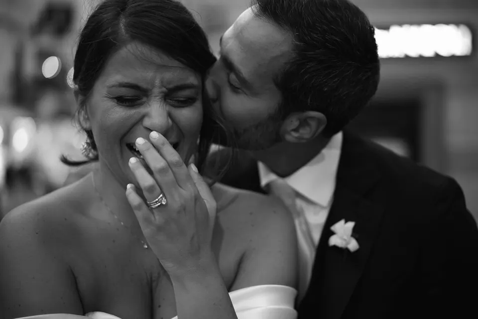 Black and white photo of the bride laughing through tears as the groom kisses her cheek.