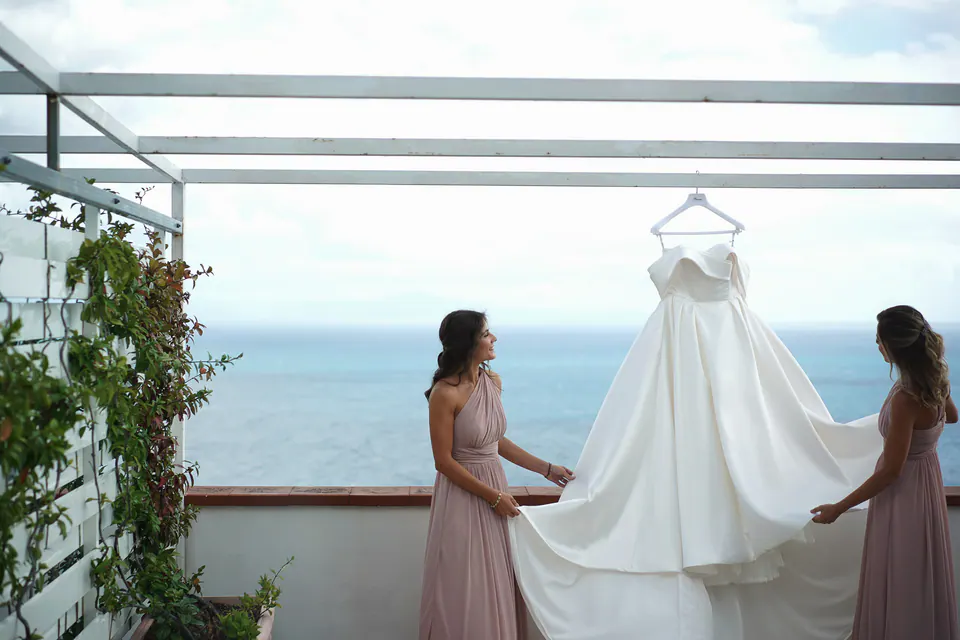 Bridesmaids holding the gown on the terrace, soft light and sea breeze creating a serene scene.