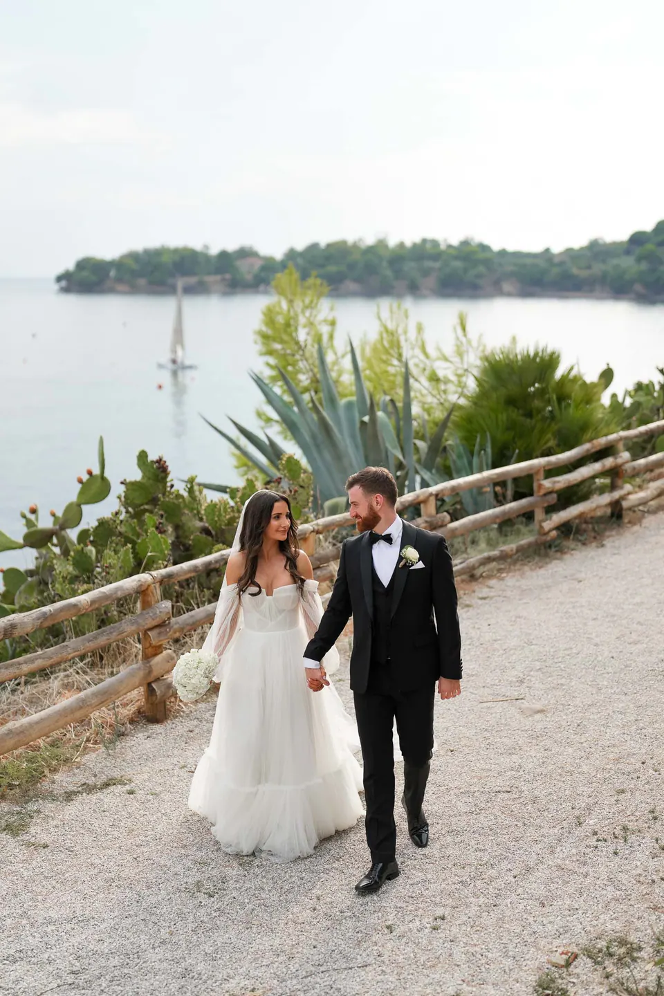Bride and groom walking on a garden path above the sea in Castellabate, an island behind them.