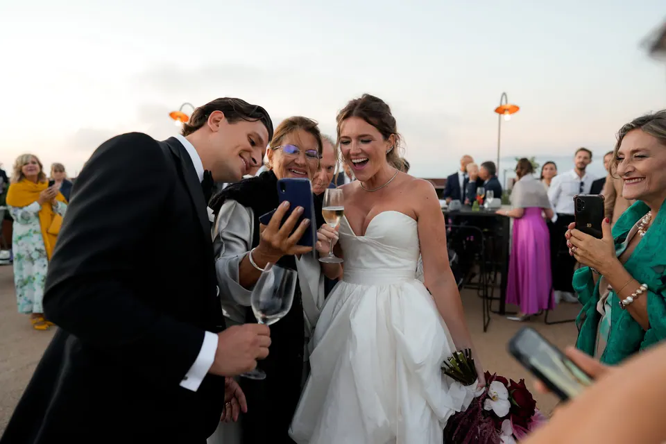 Sposi che ridono con gli invitati guardando un telefono all'aperitivo.