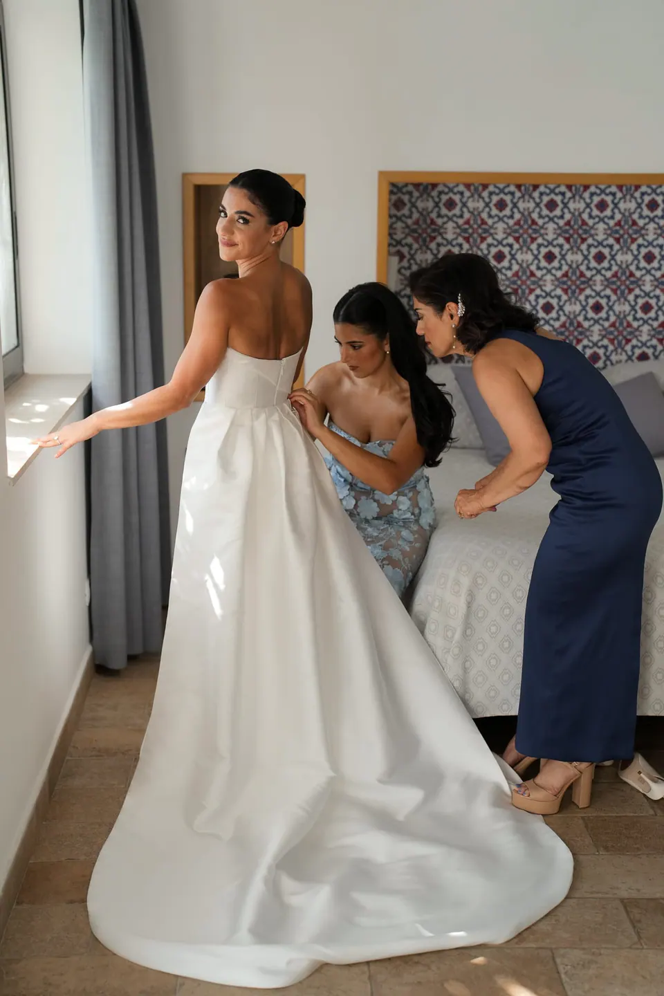 Bridesmaids fastening the bride's gown in the villa bedroom.