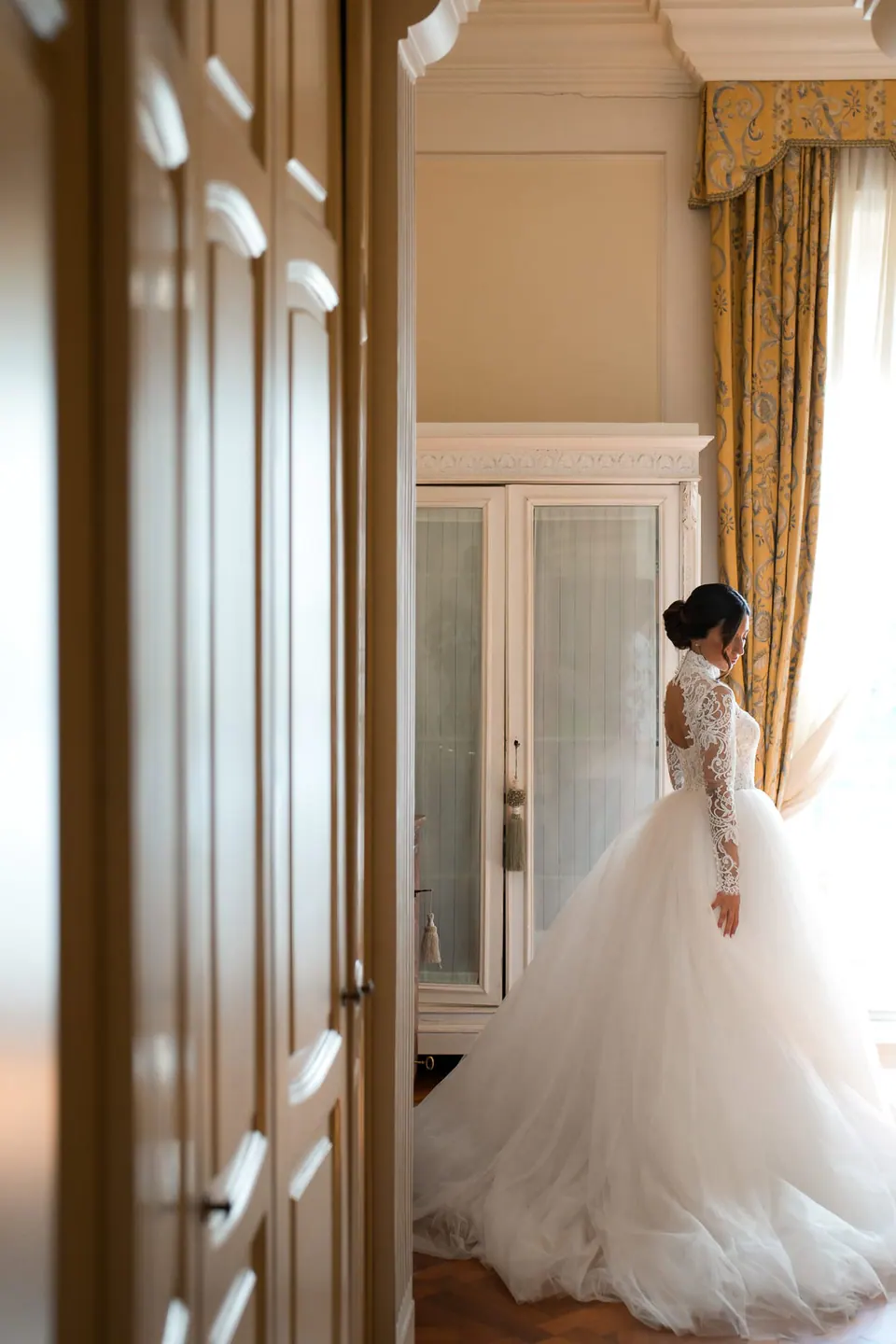 Bride by the curtains, window light on the lace of her gown.