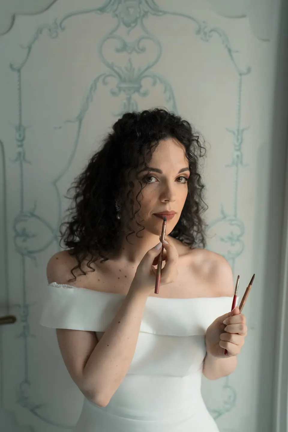 Retrato cercano de la novia sujetándose el pelo, con vestido de hombros descubiertos ante una pared pintada.