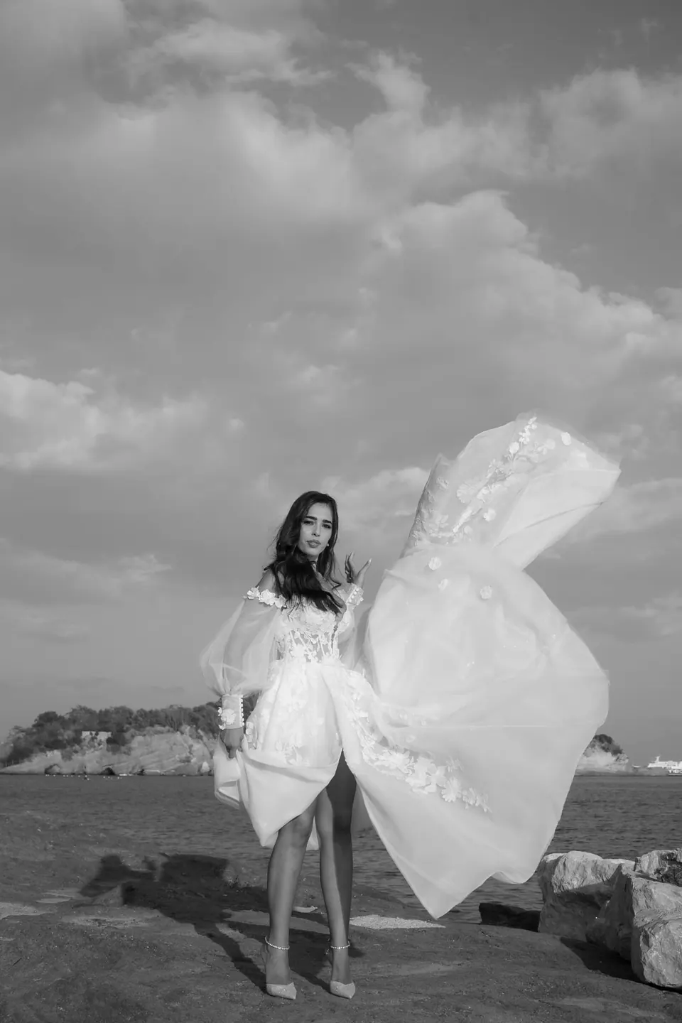 Black and white photo of the bride on the seafront, the bow of her gown lifted by the wind.