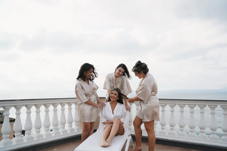 Bride laughing with her bridesmaids in matching robes on the terrace above the sea.