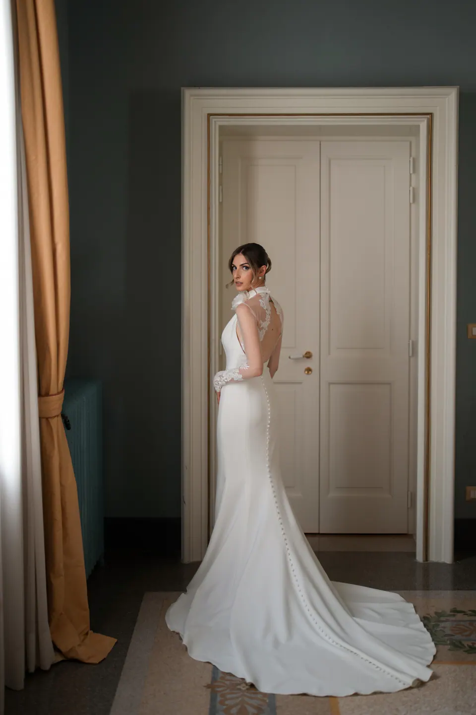 La sposa vista di spalle accanto a una porta bianca, la schiena scoperta e il lungo strascico dell'abito.