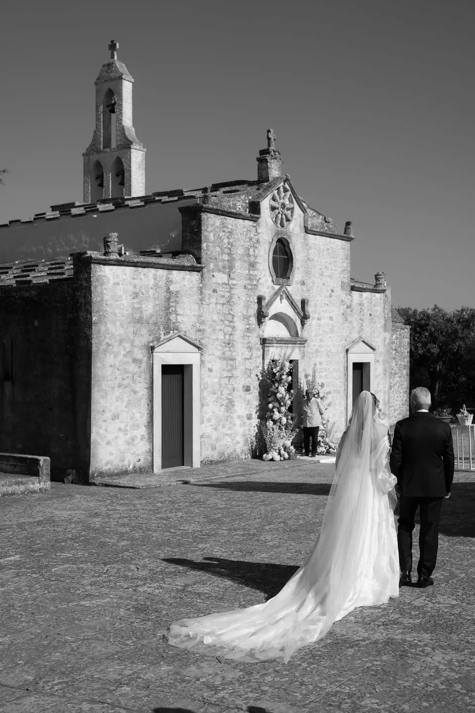 Bride standing in front of a white stone chapel, romantic Mediterranean wedding moment.