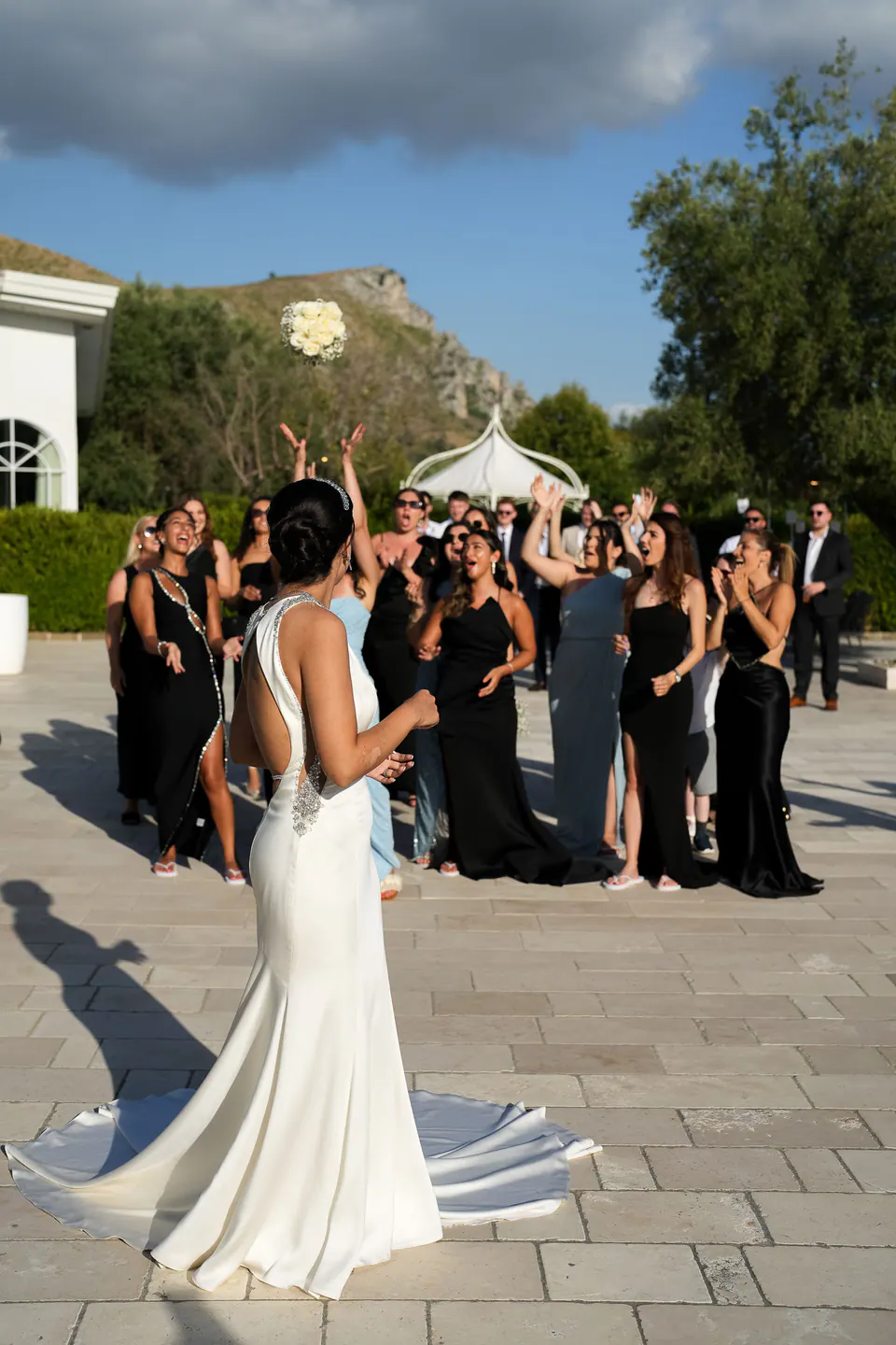 La mariée en robe de satin dos nu lance son bouquet à ses amies en robe noire.
