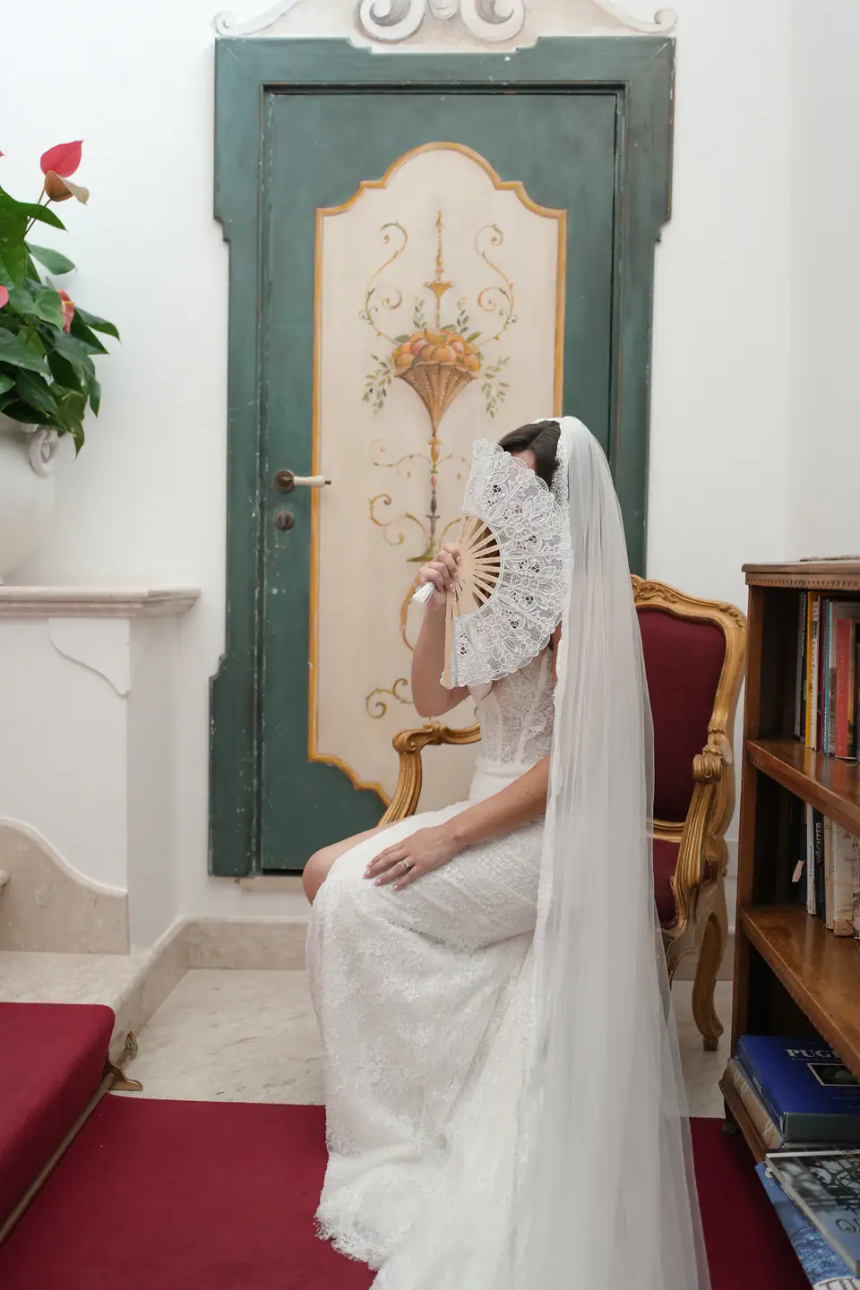 Bride sitting on a gilded chair beside a painted door, holding a lace fan.