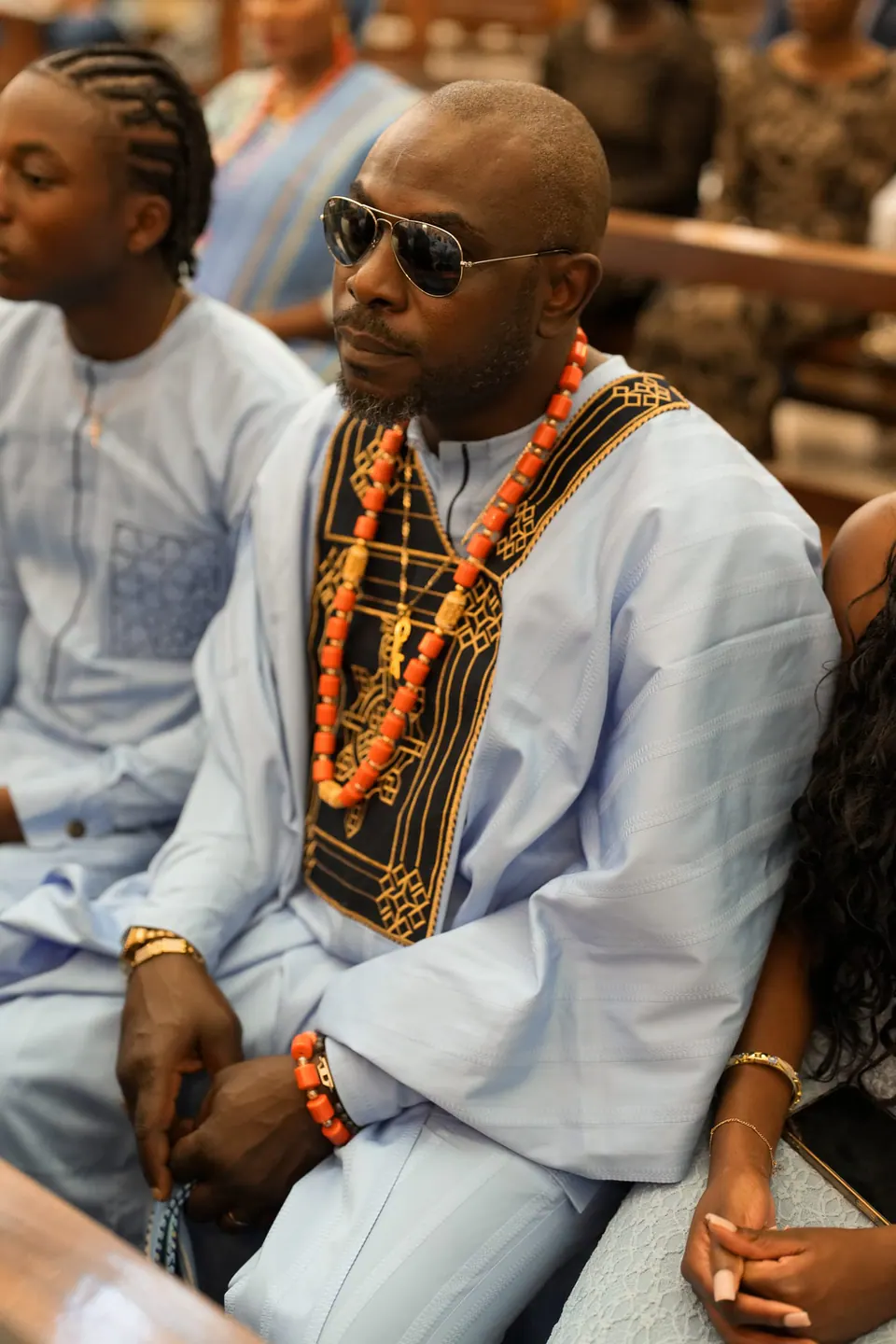 Un invitado con traje tradicional blanco, collares de coral y gafas de sol, sentado entre los invitados.