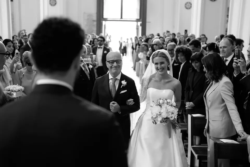 Schwarzweißfoto: Der Vater führt die Braut durch den Mittelgang zum Bräutigam.